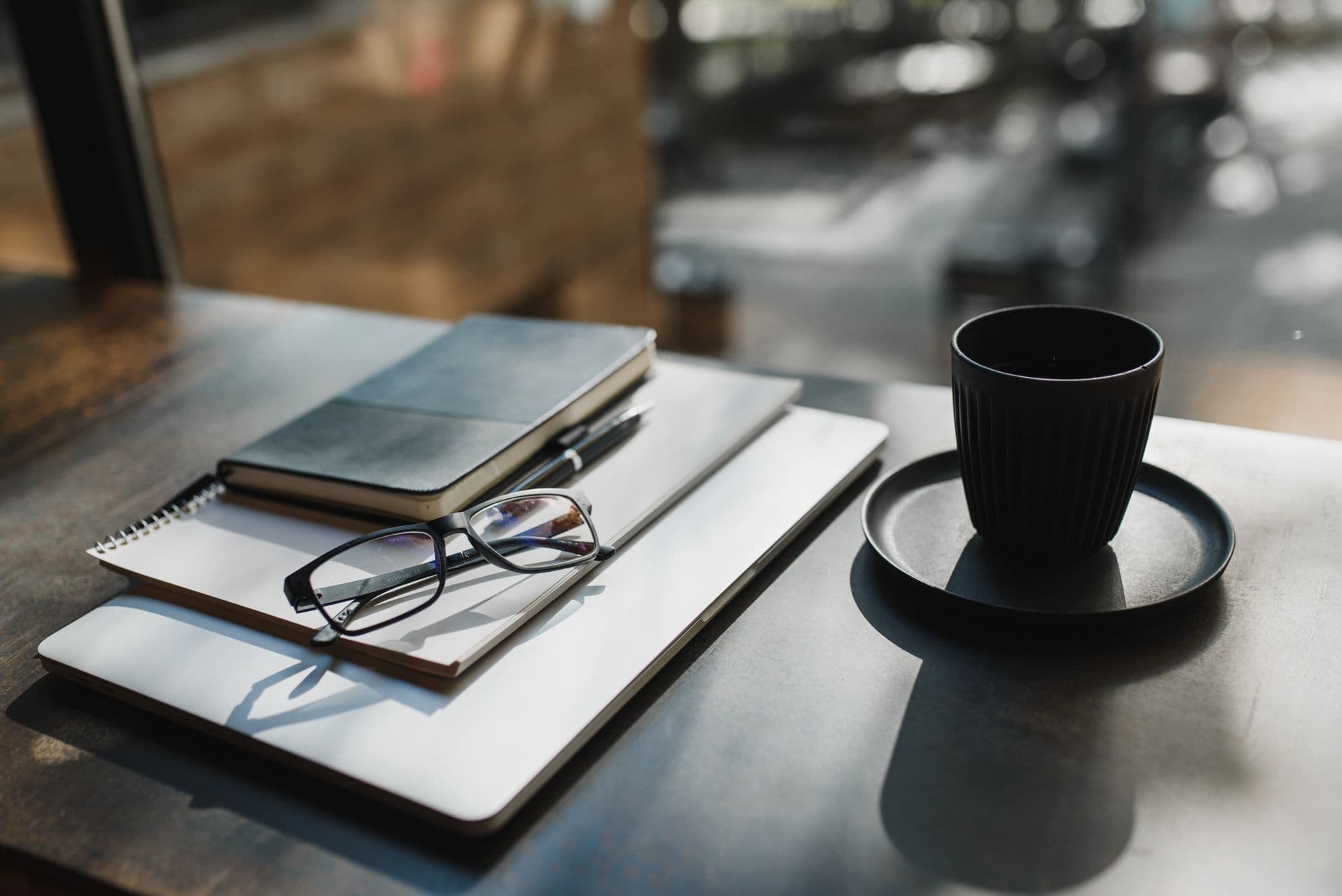Kaffekopp, briller og notatbok på et arbeidsbord ved vinduet