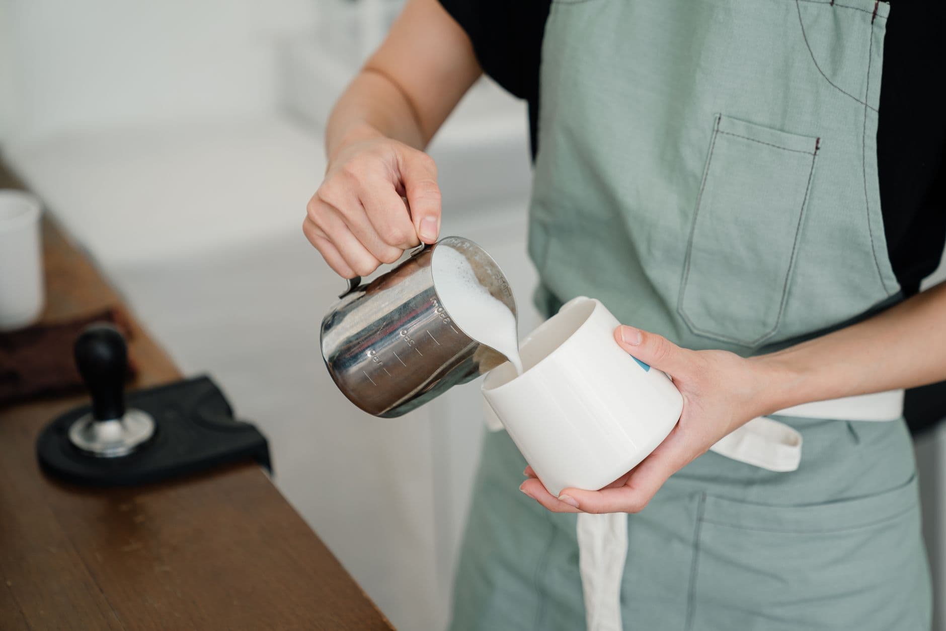 Anonym barista i grønt forkle heller melk i en hvit kopp bak kafédisken